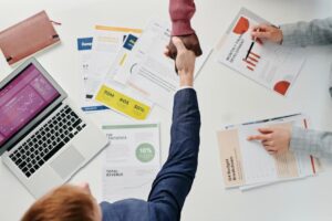 Business professionals engaging in a handshake over a desk filled with financial documents and a laptop.