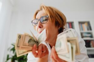 A happy woman with eyeglasses cherishing US dollar bills indoors, symbolizing wealth and financial success.
