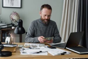 Businessman counting cash at a desk with documents and a laptop in an office.