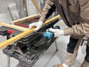 A carpenter using a power drill to work on wooden planks in an indoor workshop setting.
