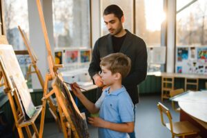 Art teacher supervises child painting in a well-lit classroom, promoting creativity.