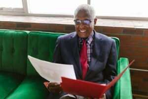 Elderly man in a blue suit sits on a green sofa, reviewing paperwork with a smile.