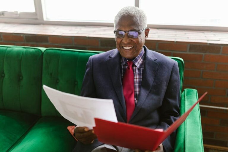 Elderly man in a blue suit sits on a green sofa, reviewing paperwork with a smile.