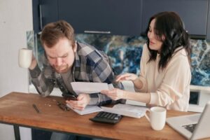 A couple sitting at a desk, reviewing bills and expenses with a calculator and laptop, looking worried.