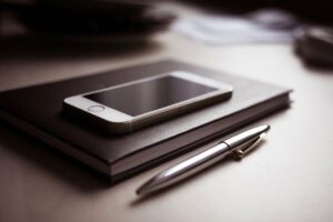 A sleek smartphone resting on a closed notebook beside a metallic pen on a desk.