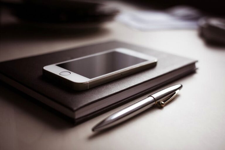 A sleek smartphone resting on a closed notebook beside a metallic pen on a desk.