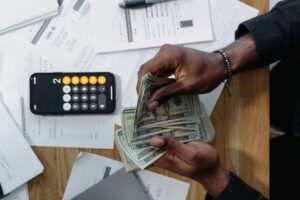 Person counting dollar bills over documents with a smartphone calculator on the desk.