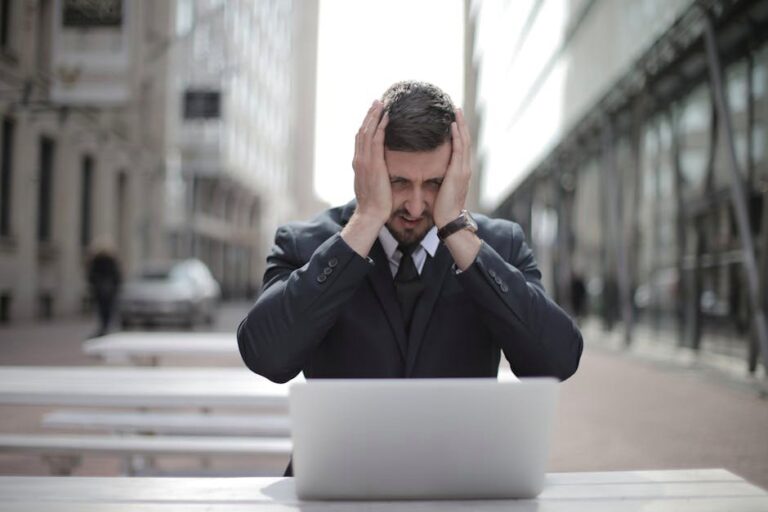 A businessman in a suit looks stressed, sitting outdoors with a laptop in a city environment.