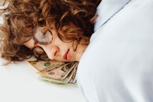 Close-up of a woman with curly hair and glasses resting her head on US dollar bills, symbolizing dreams of wealth.