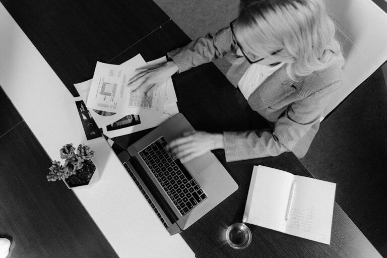 Black and white photo of a woman working at a desk with a laptop and papers.