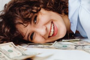 A joyful woman with curly hair lies on a pile of dollar bills, smiling brightly.