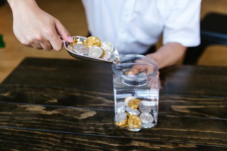 Hand placing gold and silver coins into a glass jar on a wooden table indoors.
