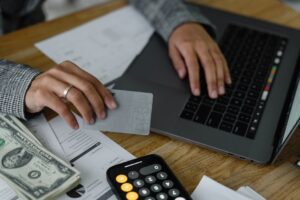 Close-up of a person using a credit card and laptop in a financial setting with cash and calculator.