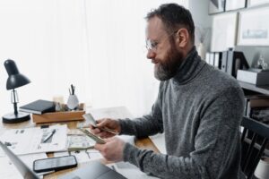 Businessman wearing glasses counting cash at office desk with documents and smartphone.