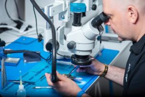 Technician examining electronic chip under a microscope in a computer lab setting.