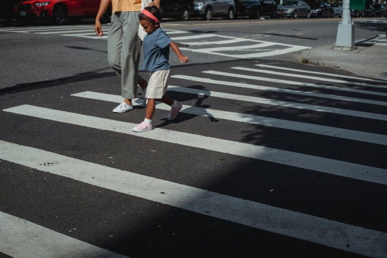 A mother and her daughter holding hands while crossing a city street on a zebra crossing.