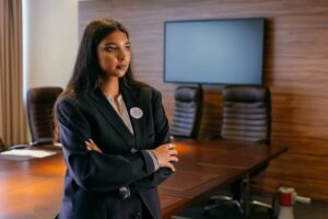 A confident woman in a business suit stands with arms crossed in a corporate boardroom.