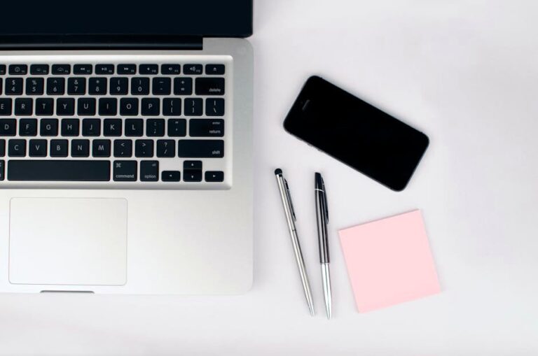 Overhead view of a sleek workspace featuring a laptop, smartphone, and stationeries on a white desk.