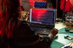 A software developer writing code on a laptop at a cluttered workbench with electronic tools.