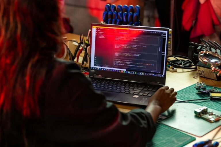 A software developer writing code on a laptop at a cluttered workbench with electronic tools.
