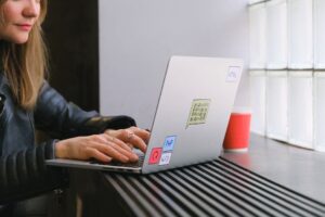 Woman using a laptop by a window in a cafe with a coffee. Ideal for tech and lifestyle themes.