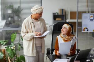 Two Muslim businesswomen in hijabs collaborating at office desk, engaged in discussion.