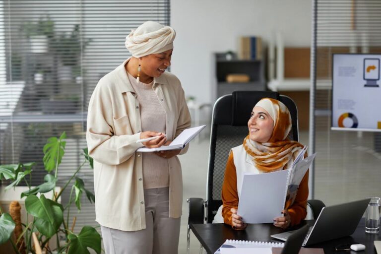 Two Muslim businesswomen in hijabs collaborating at office desk, engaged in discussion.