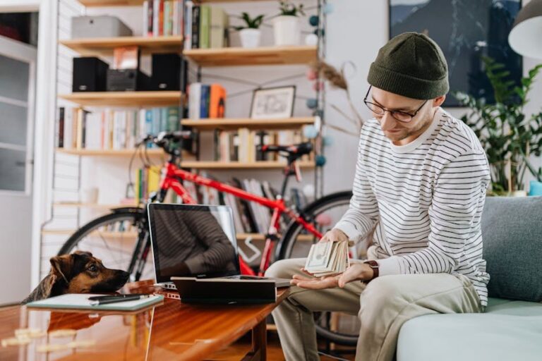 Young man counting cash at home with his laptop, wearing a beanie and glasses, in a casual living room.