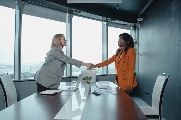 Two businesswomen shaking hands in a modern office setting, symbolizing partnership and agreement.