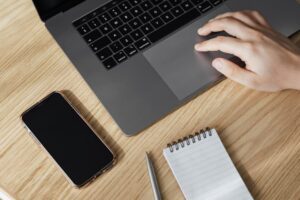 Aerial view of a hand on a laptop trackpad, with a smartphone and notepad on a wooden desk.