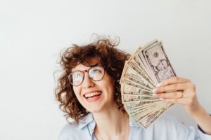 Happy woman with curly hair and glasses holding US dollar bills against a white background.
