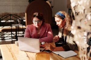 Two women working together on a laptop in a cozy café setting.