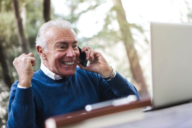 Elderly man smiling and cheering while talking on the phone using laptop outdoors.