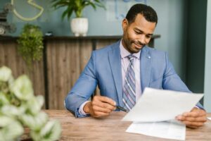 Smiling businessman in blue suit reviewing documents at a wooden table in modern office setting.