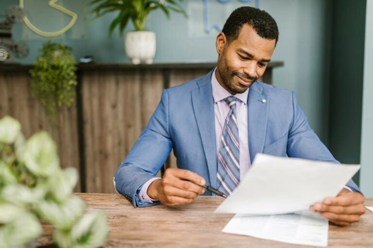 Smiling businessman in blue suit reviewing documents at a wooden table in modern office setting.