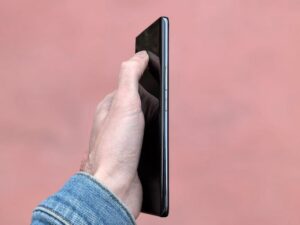 A hand in a denim jacket holds a sleek smartphone against a soft pink background.