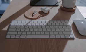 Sleek workspace setup featuring a white keyboard, mouse, and wireless earbuds on a wooden table with natural lighting.