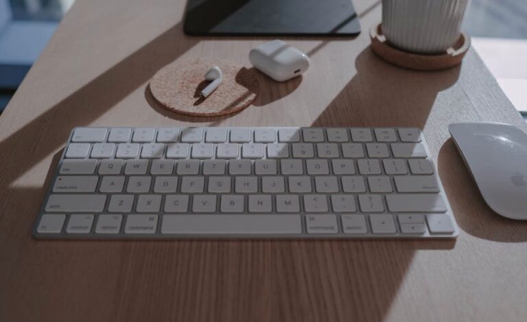 Sleek workspace setup featuring a white keyboard, mouse, and wireless earbuds on a wooden table with natural lighting.