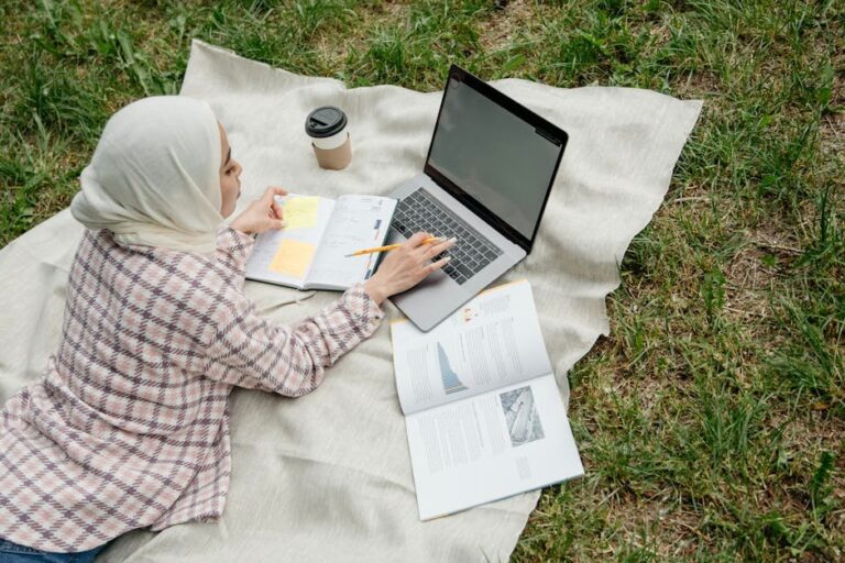 Muslim woman using laptop and notebook in park, engaging in remote work.