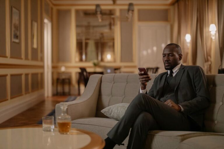 Elegant businessman using smartphone while relaxing in hotel lounge with whiskey on the table.