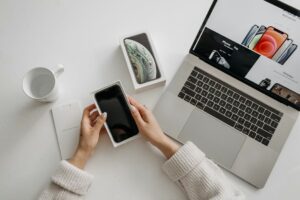 Flat lay of hands unboxing smartphone next to laptop on a white desk, representing modern digital lifestyle.