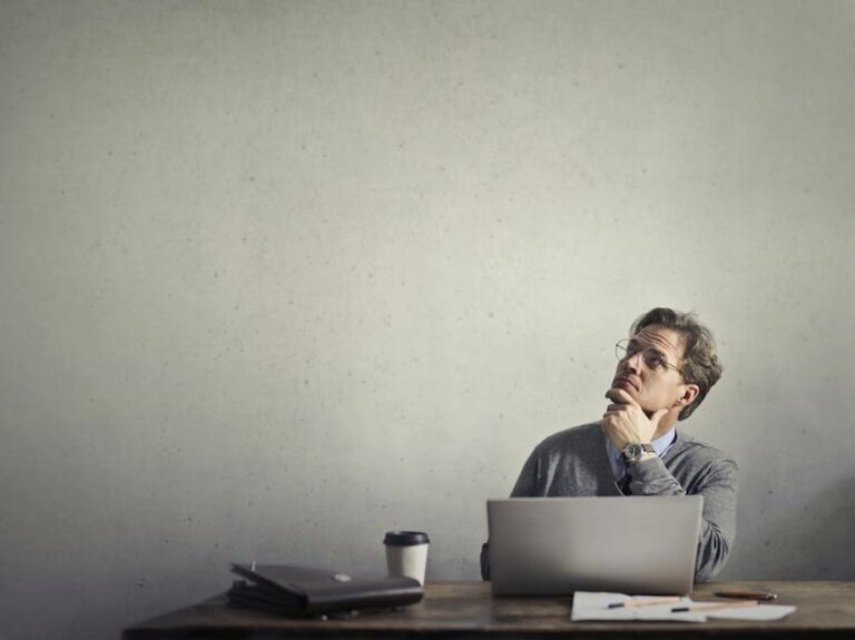 Thoughtful businessman looking up while working at his desk with a laptop and coffee.