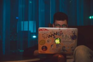 A man focused on working on his laptop with stickers at night, indoors.