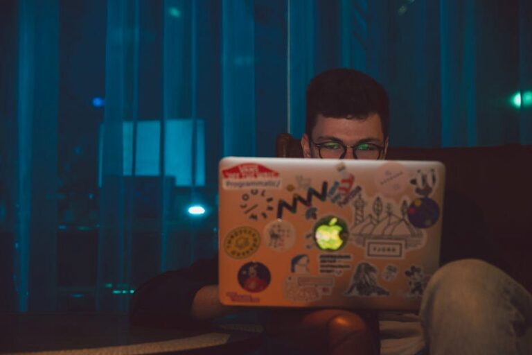 A man focused on working on his laptop with stickers at night, indoors.