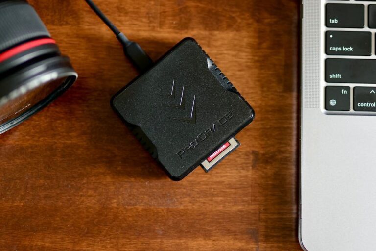 Close-up shot of a card reader with a memory card on a wooden surface alongside a camera lens and laptop.