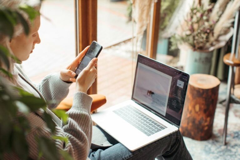 A woman using her smartphone and laptop for remote work indoors, capturing a cozy workspace.