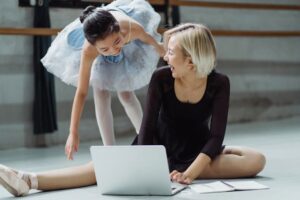 A young ballerina and her teacher enjoy a moment of laughter while using a laptop in a dance studio.