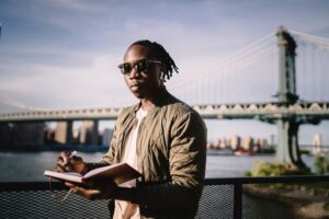 A young man with sunglasses writing in a notebook by a famous city bridge under sunny skies.
